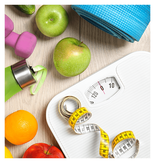 A variety of fitness and health items on a wooden surface, including green and red apples, an orange, a pink dumbbell, a green water bottle, a blue yoga mat, a white scale with a yellow measuring tape, and a purple-handled item.