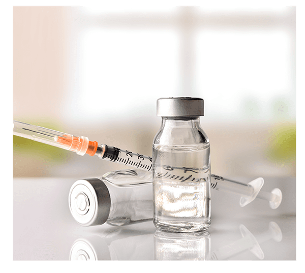 Two small glass medicine bottles and a syringe with a needle, set on a white surface with a blurred bright background.
