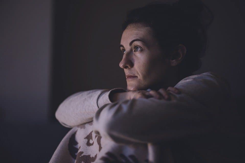 A woman with dark hair resting her chin on her crossed arms, looking thoughtfully into the distance in dim lighting.